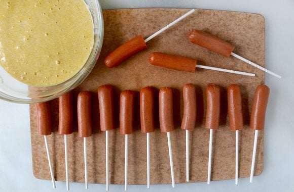 Cutting board with row of hot dogs on sticks next to bowl with batter