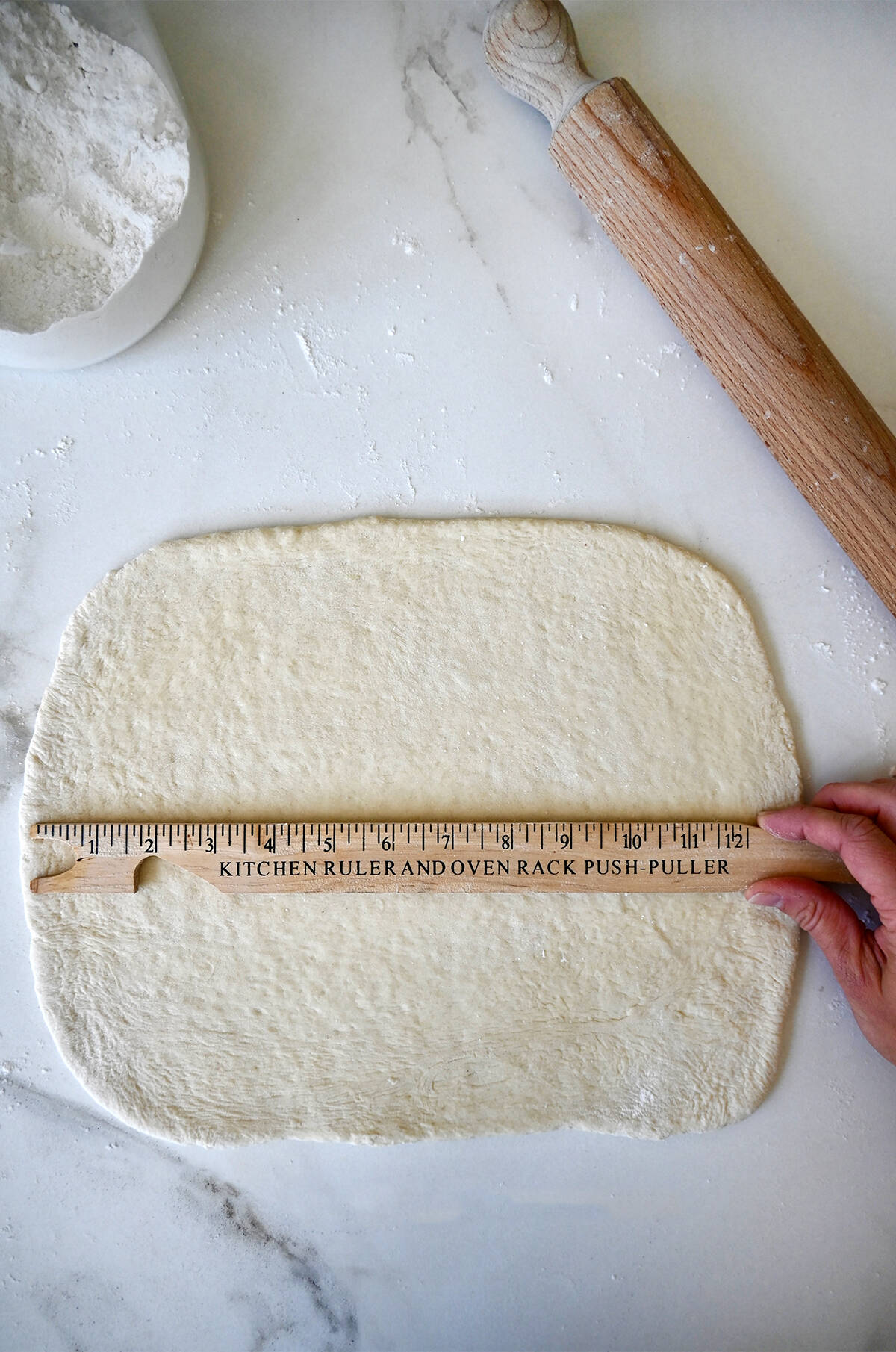 A hand holding a ruler over pizza dough rolled into a 13'' rectangle. 