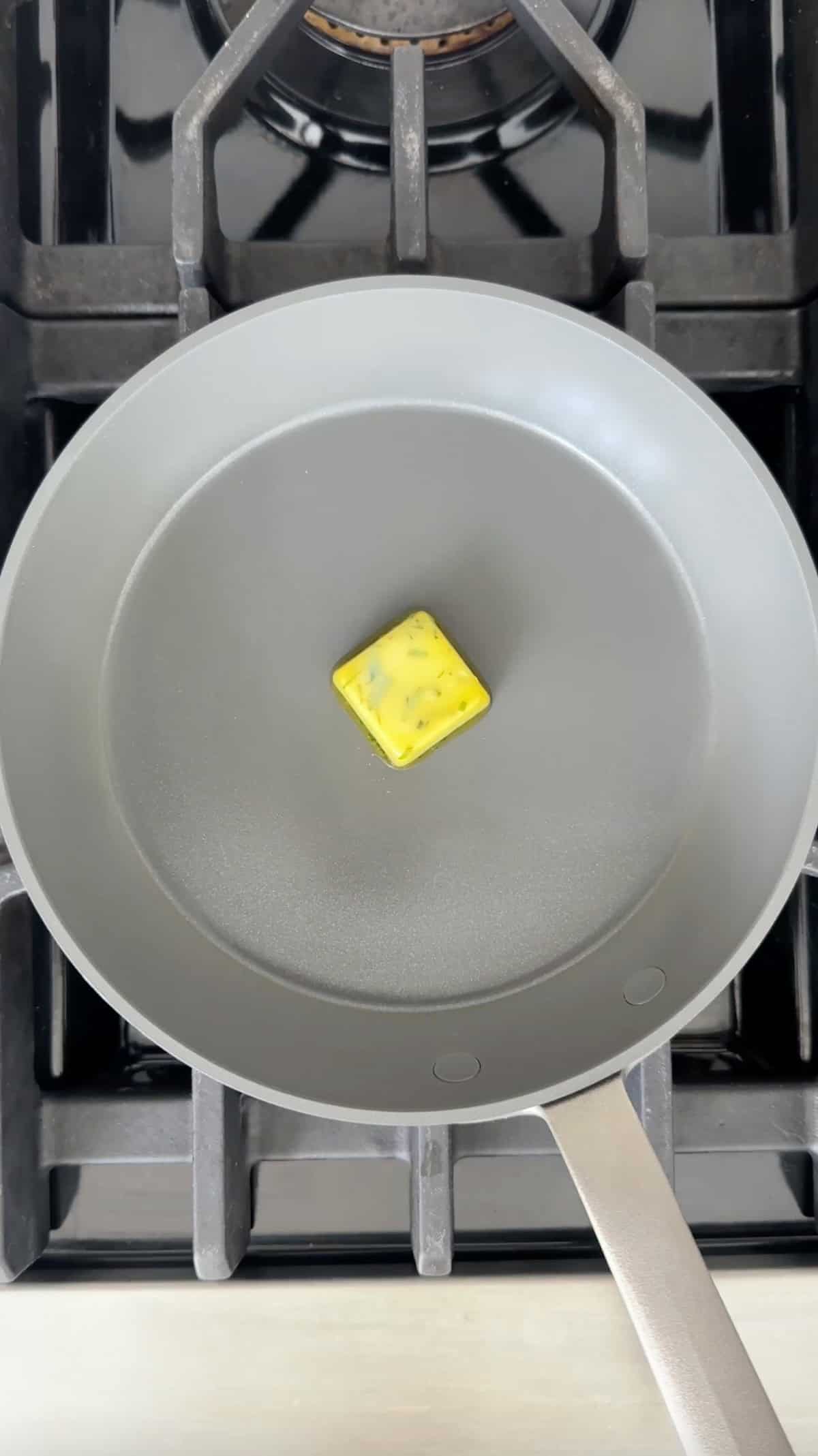 A frozen fresh herb olive oil cube melting in a skillet on a stovetop.