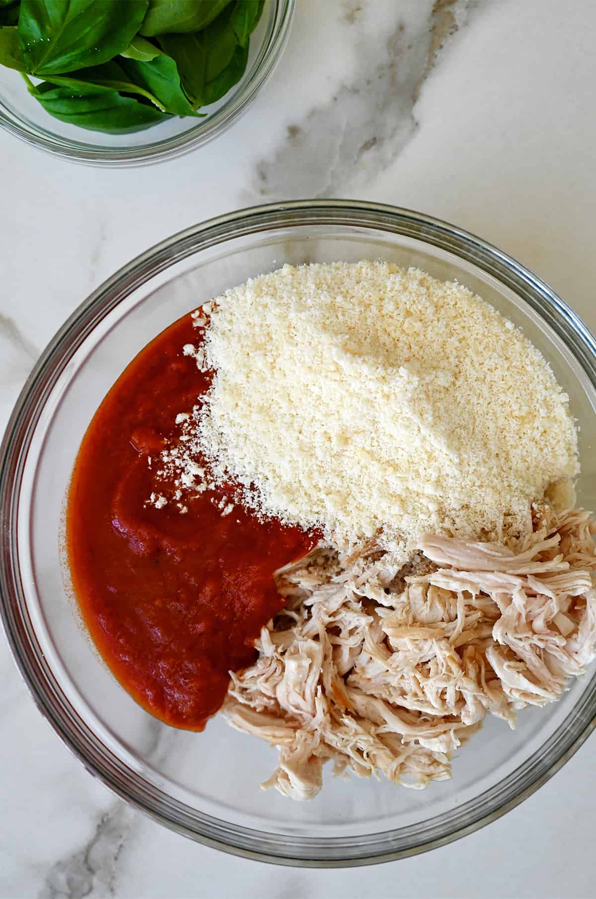A glass bowl containing shredded chicken, marinara sauce and grated Parmesan cheese.