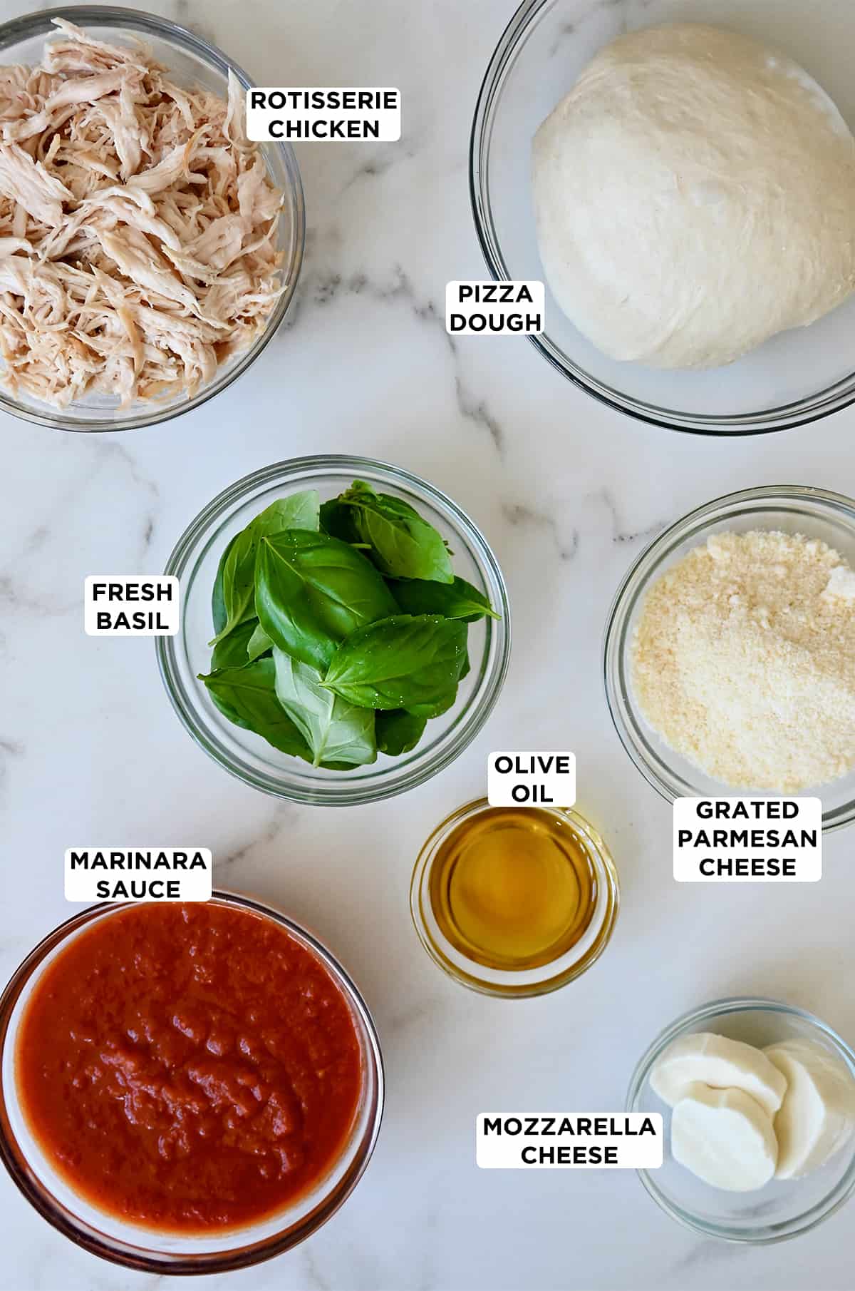 Various sizes of glass bowls containing shredded chicken, pizza dough, fresh basil, olive oil, grated parmesan cheese, mozzarella cheese and marinara sauce.
