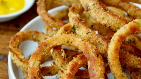 A white plate containing baked onion rings with bowls of ketchup and mustard in the background