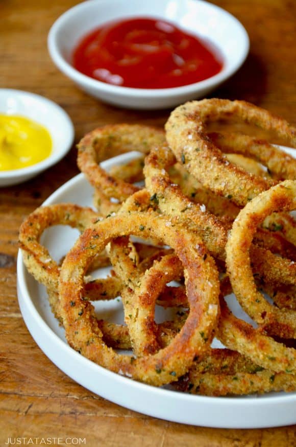 A white plate containing baked onion rings with bowls of ketchup and mustard in the background