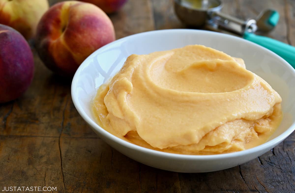 A white bowl containing creamy peach frozen yogurt next to a few fresh peaches.