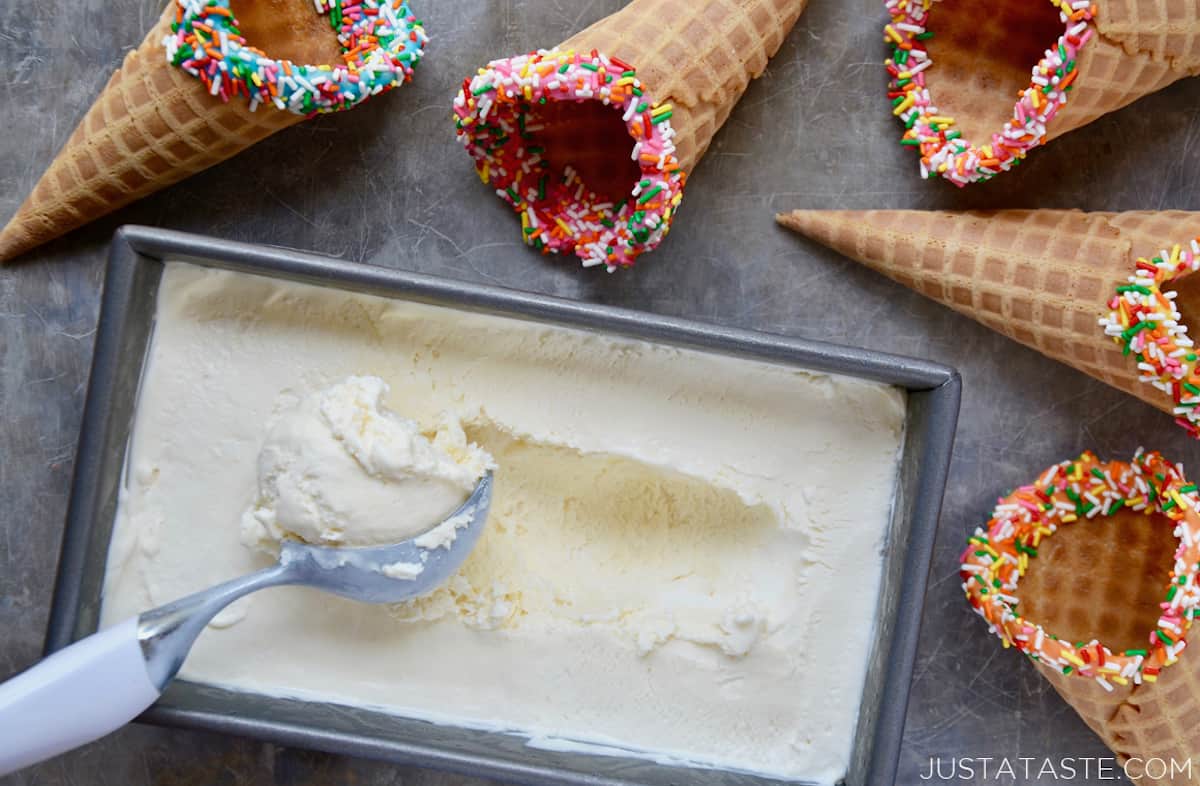 A top-down view of a loaf pan containing no-churn vanilla ice cream with an ice cream scoop next to waffle cones with rainbow sprinkles.