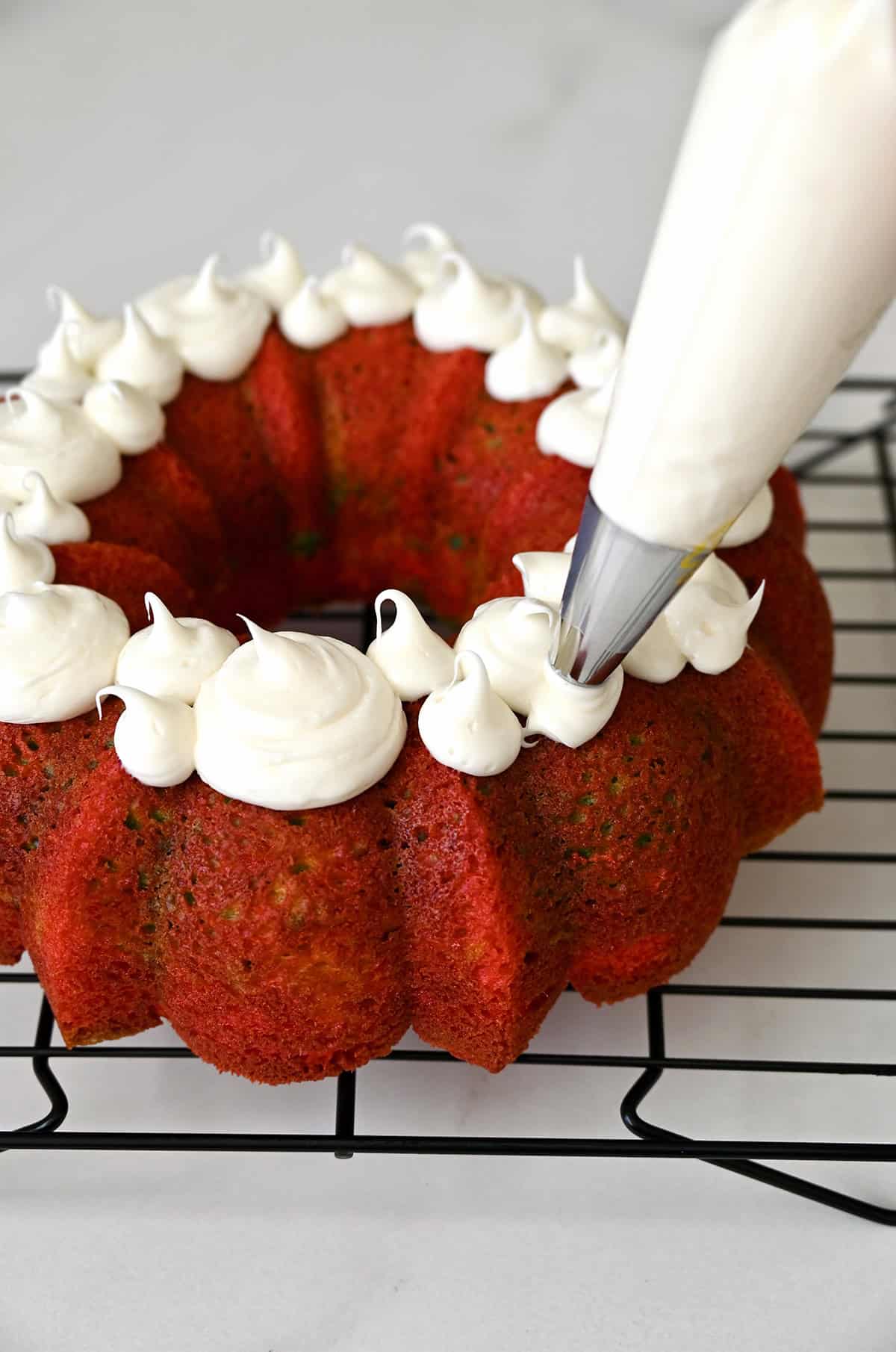 Piping vanilla frosting onto a rainbow cake on a wire cooling rack.
