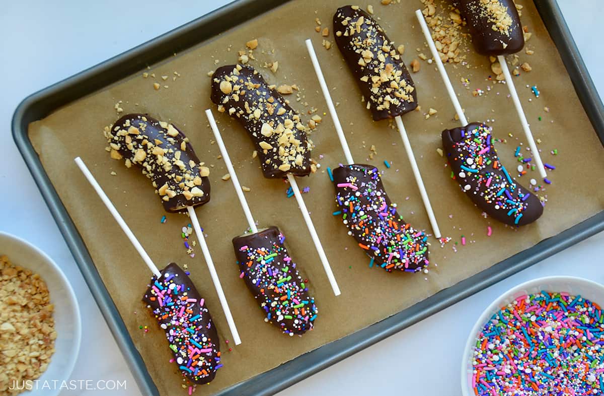 Chocolate-covered frozen bananas with sticks on top of a parchment paper-lined baking sheet.