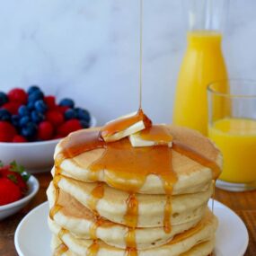 Stack of Light and Fluffy Buttermilk Pancakes topped with butter and maple syrup with orange juice and fruit in background