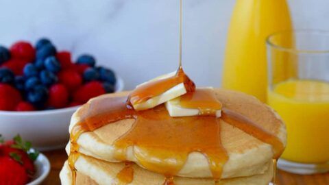 Stack of Light and Fluffy Buttermilk Pancakes topped with butter and maple syrup with orange juice and fruit in background