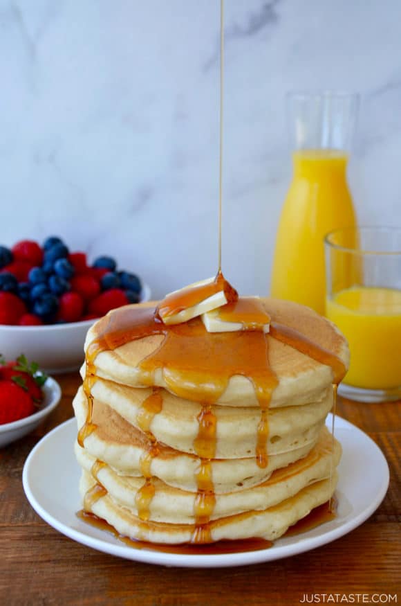 Stack of Light and Fluffy Buttermilk Pancakes topped with butter and maple syrup with orange juice and fruit in background