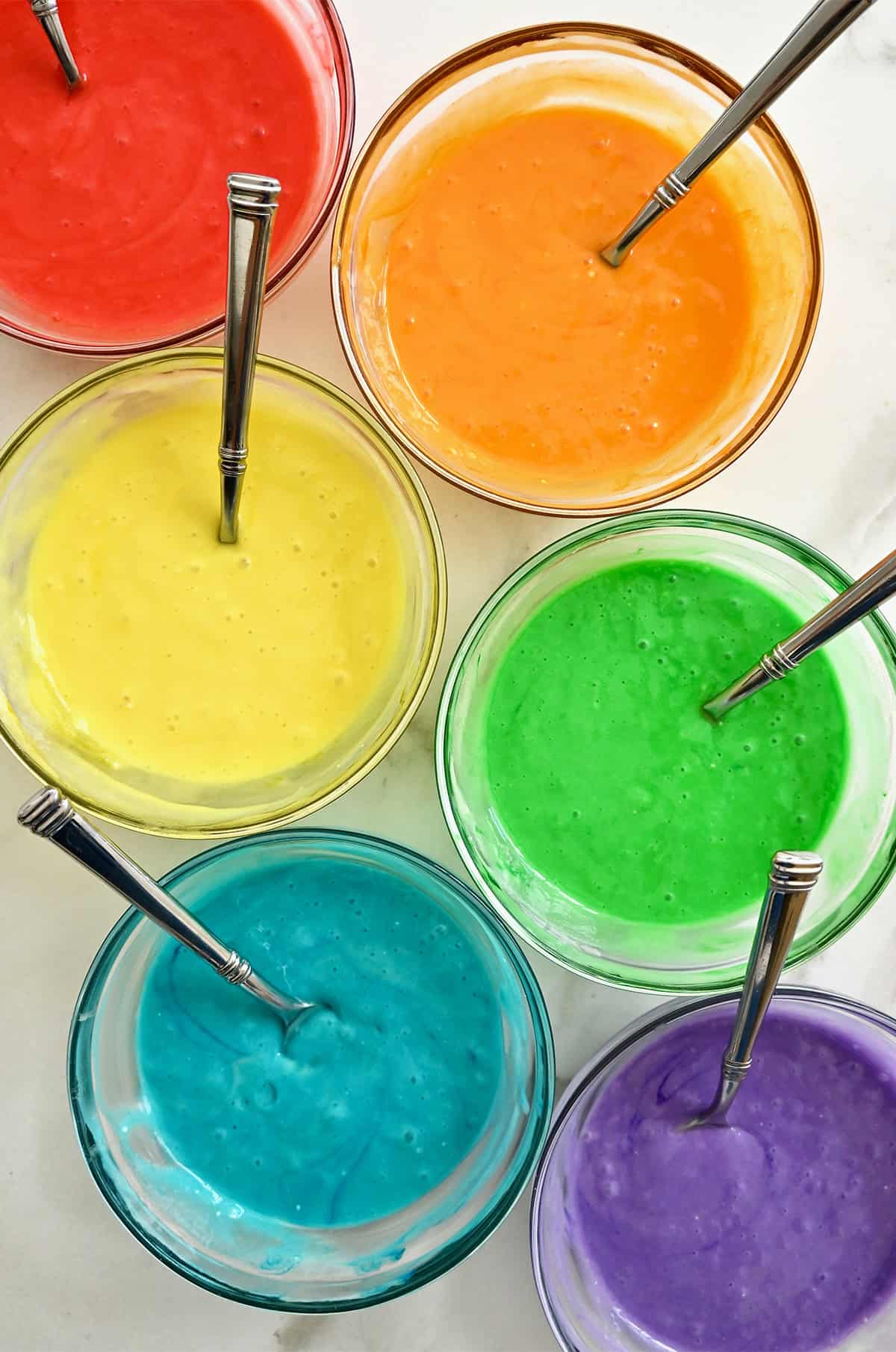 Six bowls containing cake batter dyed the colors of the rainbow: red, orange, yellow, green, blue and purple.