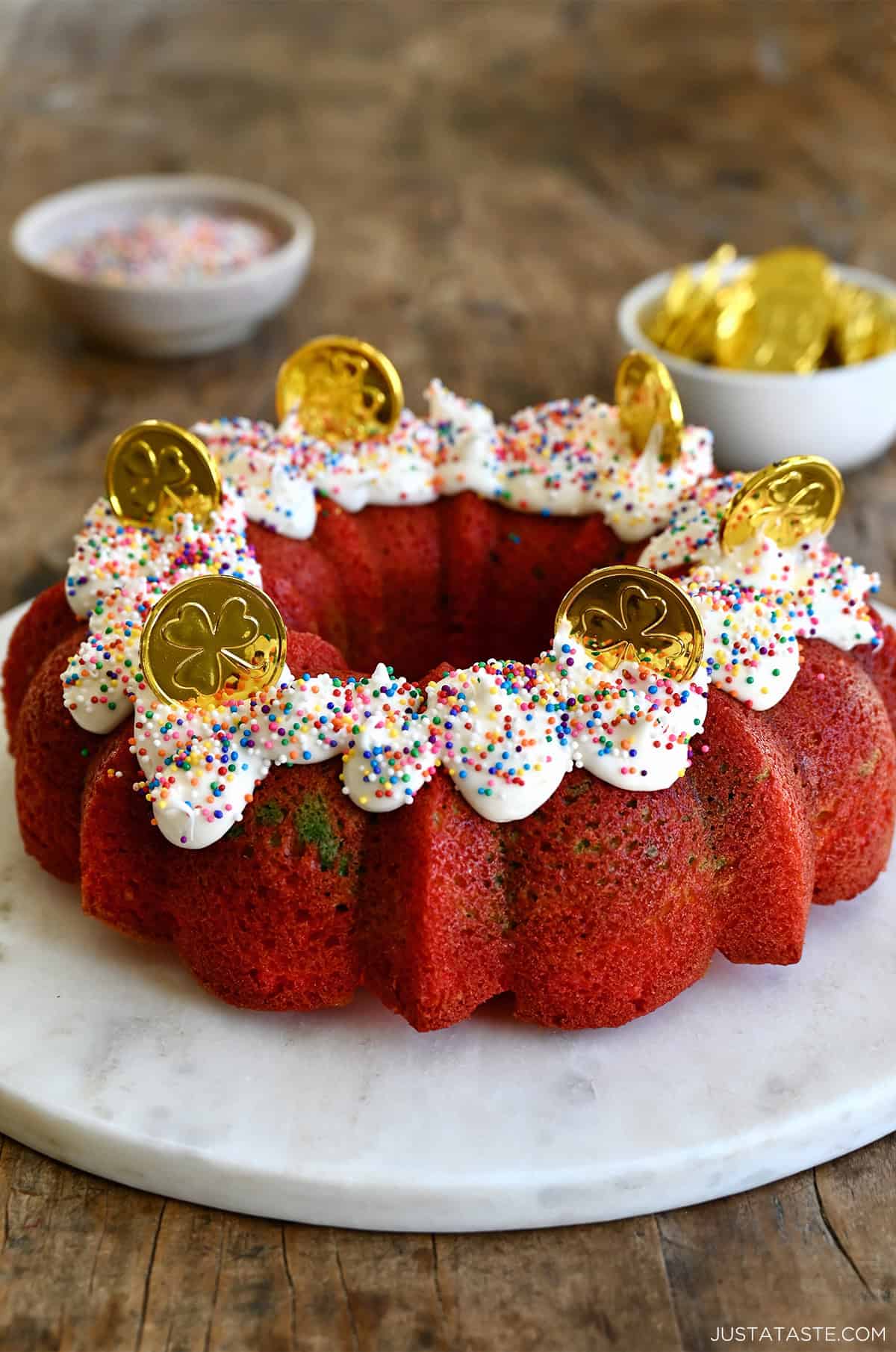 Rainbow cake topped with vanilla frosting, rainbow sprinkles and milk chocolate coins on a round cake platter.