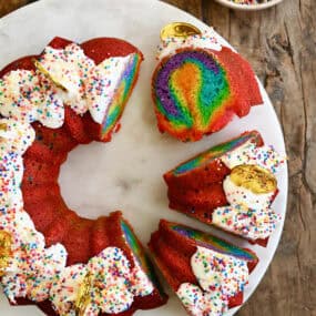 Slices of rainbow cake with vanilla frosting and rainbow sprinkles on a cake serving plate.