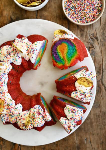 Slices of rainbow cake with vanilla frosting and rainbow sprinkles on a cake serving plate.