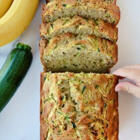 A sliced loaf of Zucchini Banana Bread with a child's hand touching it