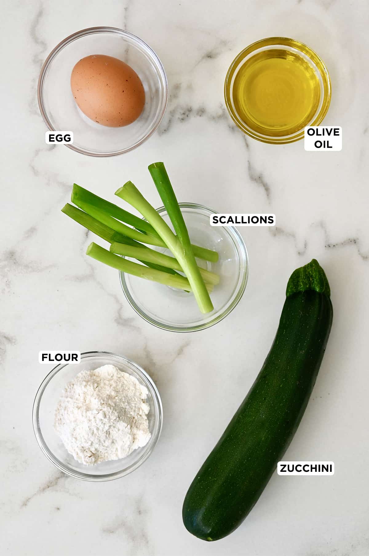 A large zucchini next to clear bowls containing an egg, olive oil, scallions and flour.