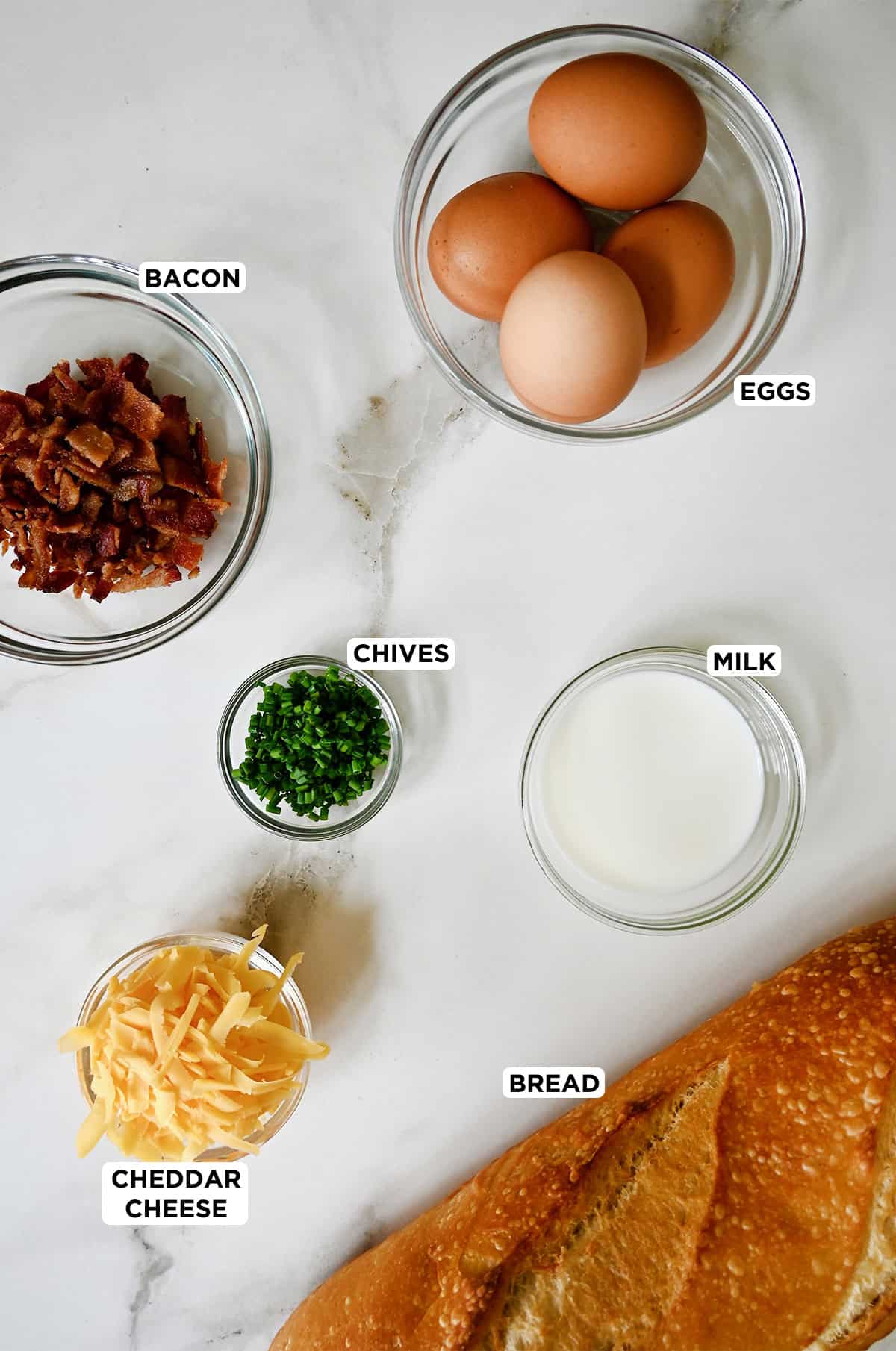 A loaf of sourdough bread next to small bowls containing shredded cheddar cheese, chopped chives, cooked crumbled bacon, eggs and milk.