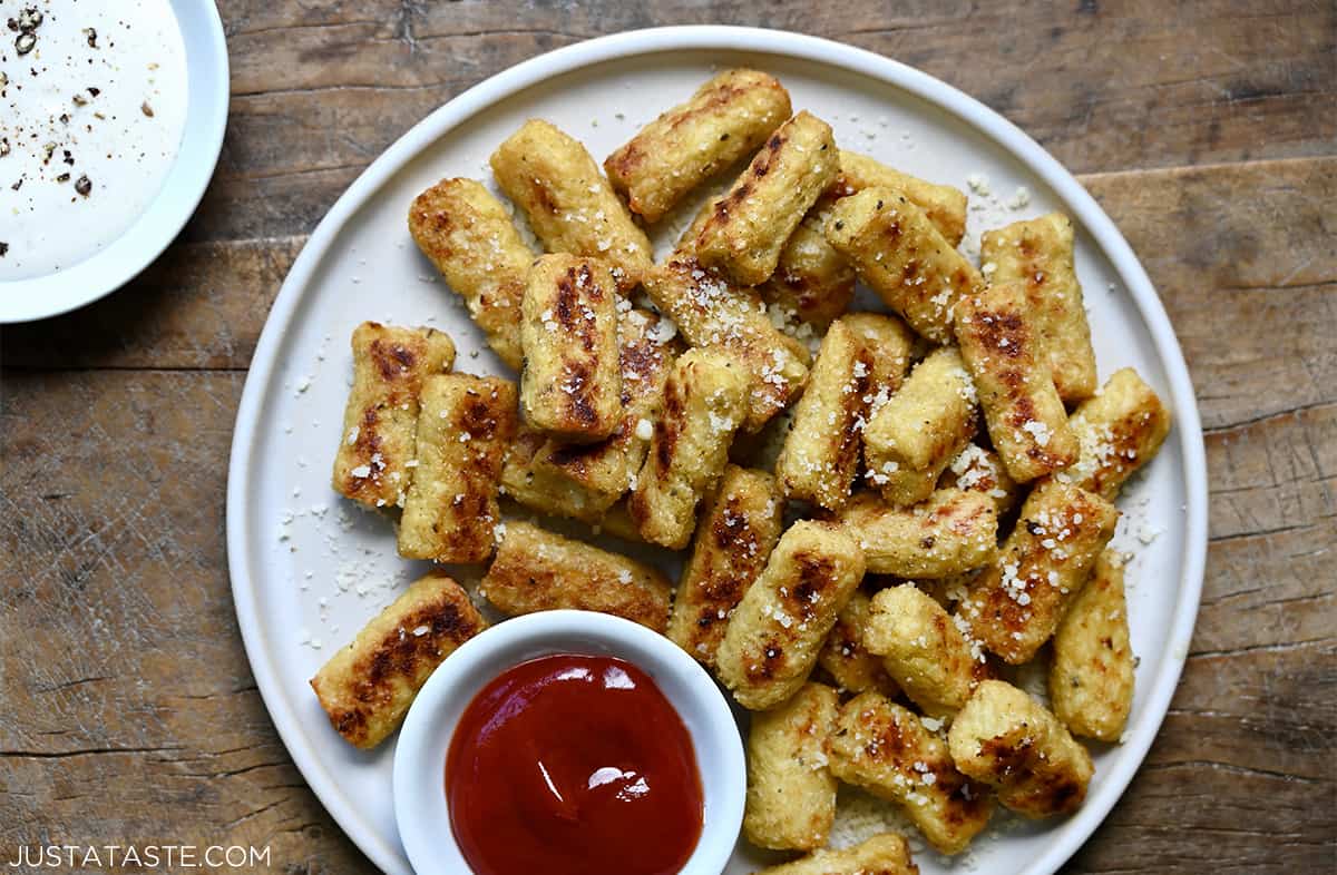 Cauliflower tots piled high on a white dinner plate surrounding a small bowl filled with ketchup.