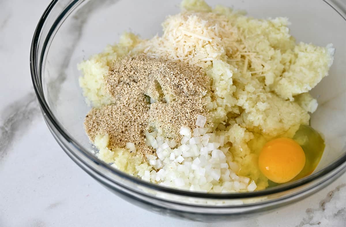 A clear bowl containing cauliflower rice, grated Parmesan cheese, breadcrumbs, diced onion and an egg.