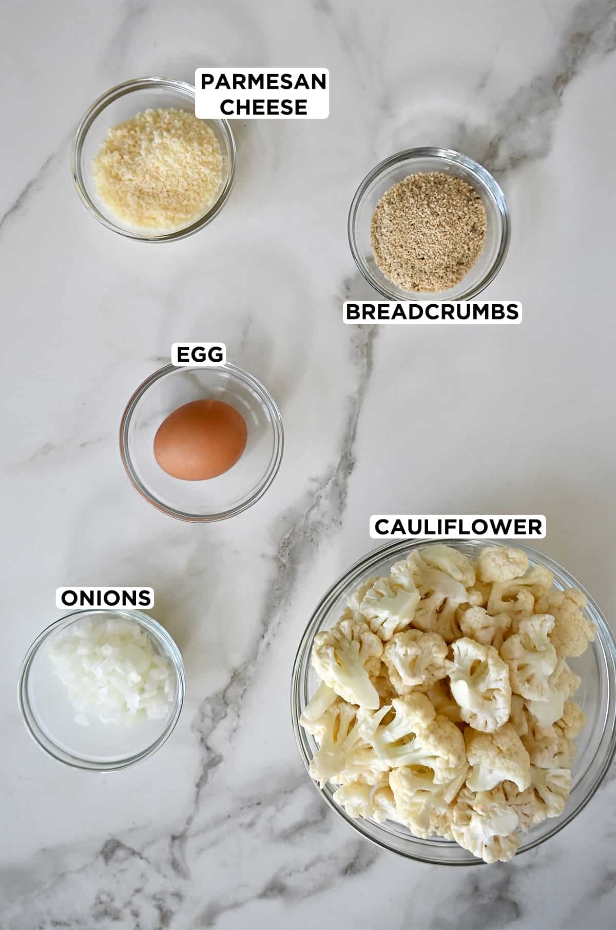 Various sizes of clear glass bowls containing grated Parmesan cheese, breadcrumbs, cauliflower florets, diced white onions and an egg.
