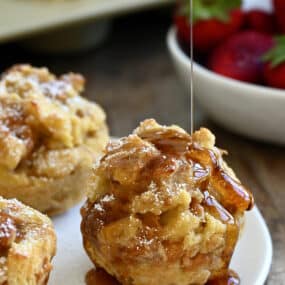 Maple syrup being drizzled atop a cinnamon French toast muffin on a white plate with two more French toast cups.