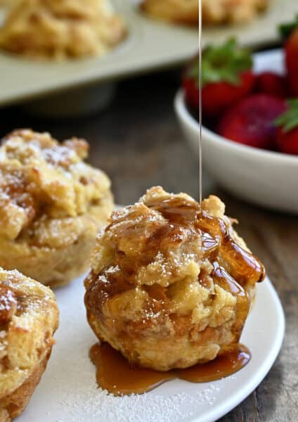 Maple syrup being drizzled atop a cinnamon French toast muffin on a white plate with two more French toast cups.