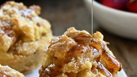 Maple syrup being drizzled atop a cinnamon French toast muffin on a white plate with two more French toast cups.