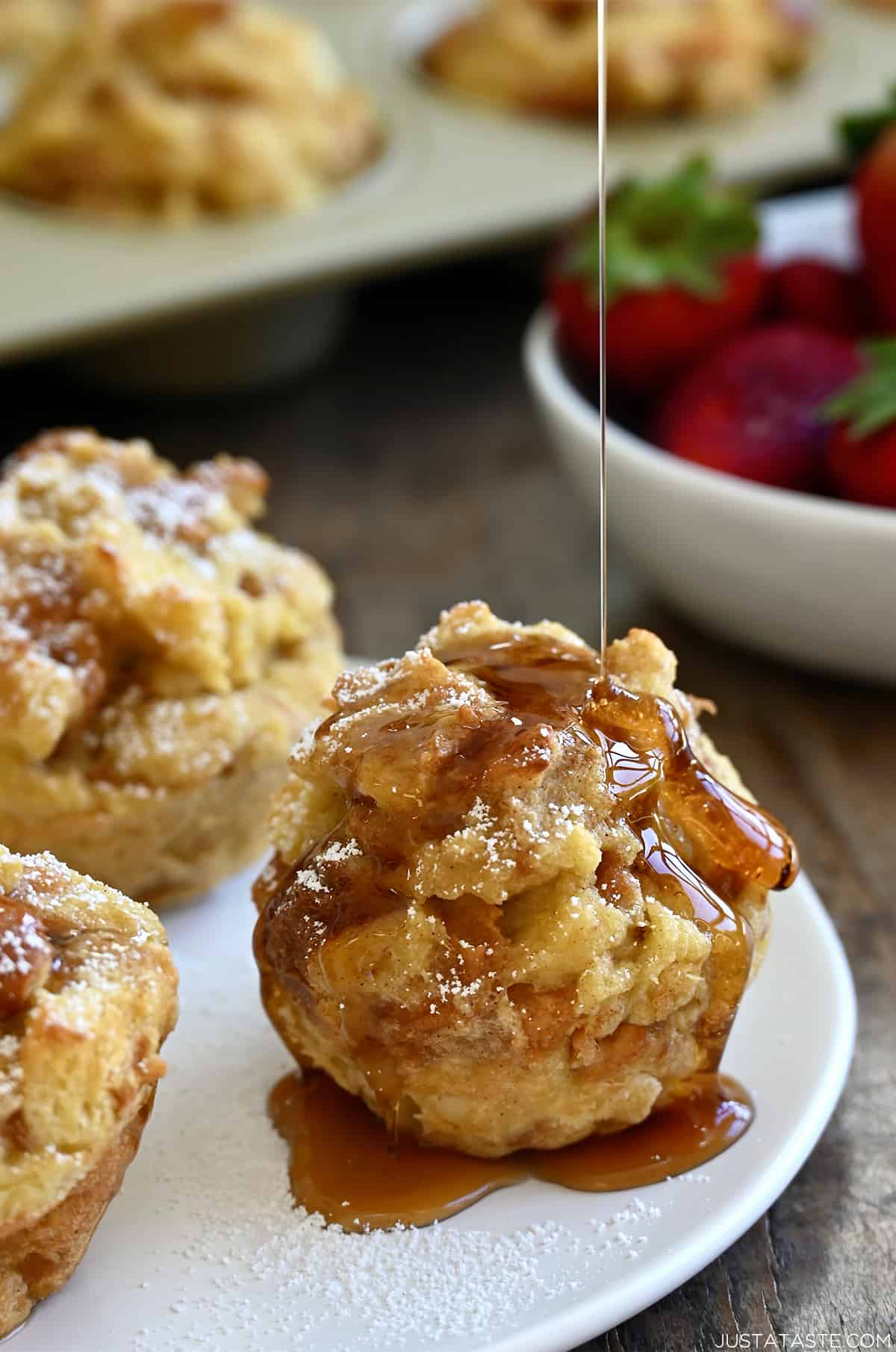 Maple syrup being drizzled atop a cinnamon French toast muffin on a white plate with two more French toast cups.