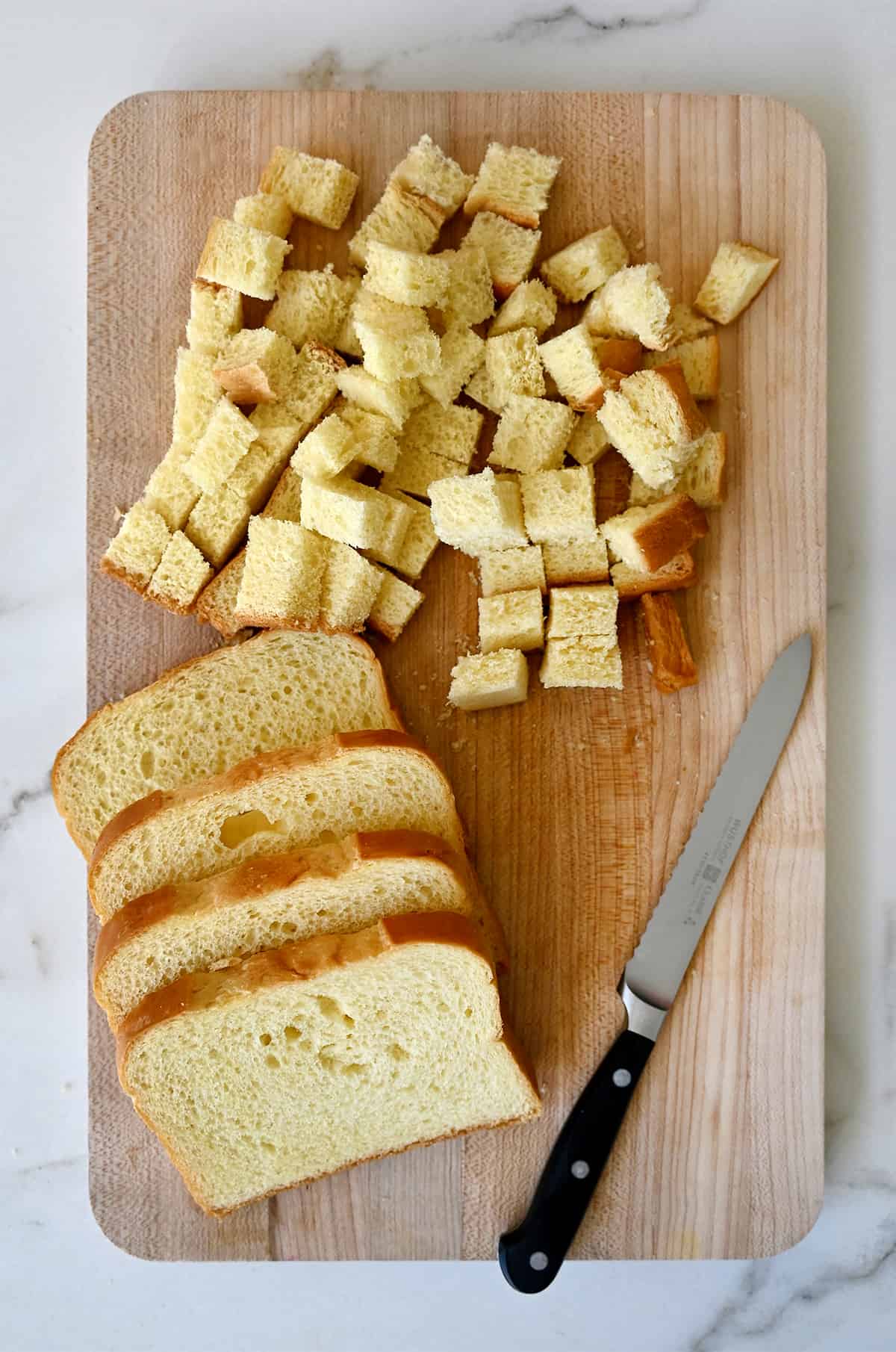 Diced brioche next to four slices of brioche and a sharp knife on a wood cutting board.