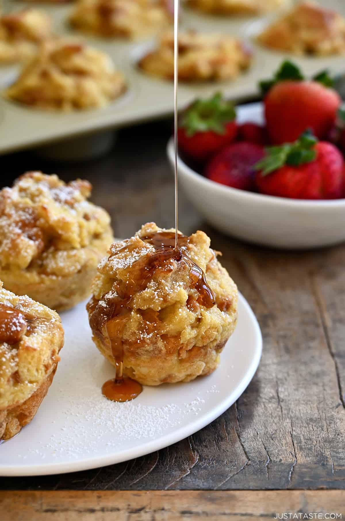 A French toast muffin on a white plate being drizzled with maple syrup. A bowl of fresh strawberries and a muffin tin are nearby.