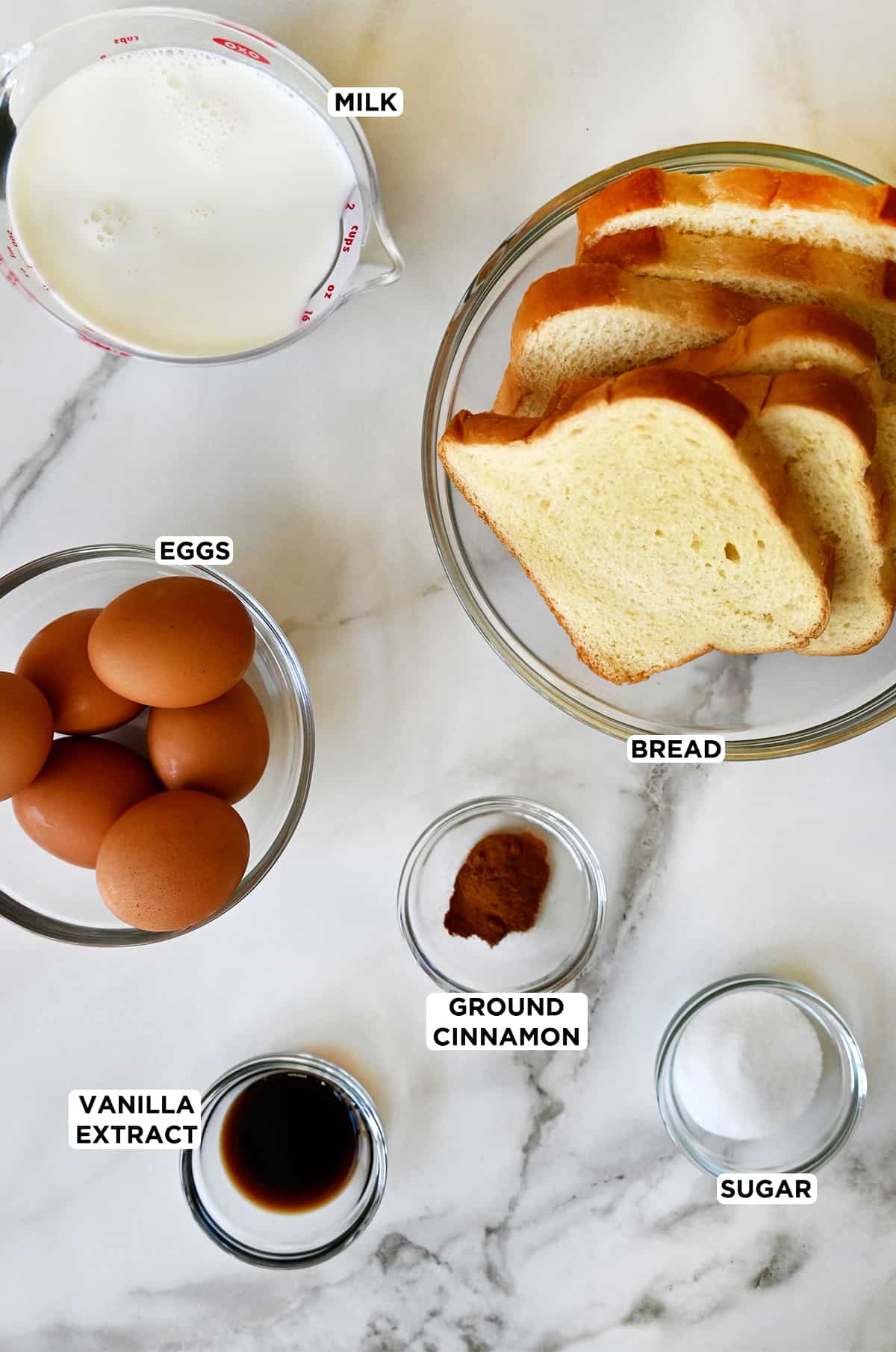 Various sizes of clear bowls containing the ingredients needed to make French toast muffins, including Brioche bread, ground cinnamon, sugar, vanilla extract and whole milk.