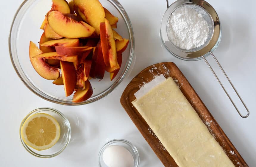 Folded puff pastry next to a bowl of fresh peach slices, powdered sugar, a halved lemon and a single egg.