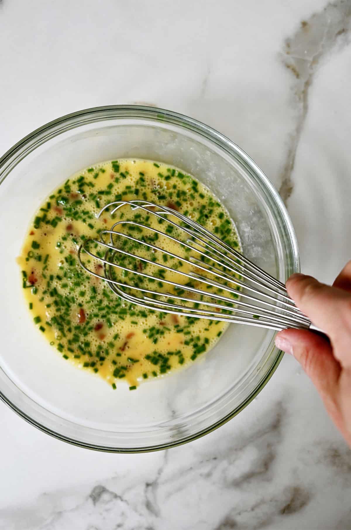 A hand holding a whisk into a bowl with eggs being whisked together with milk, cheese and bacon pieces.