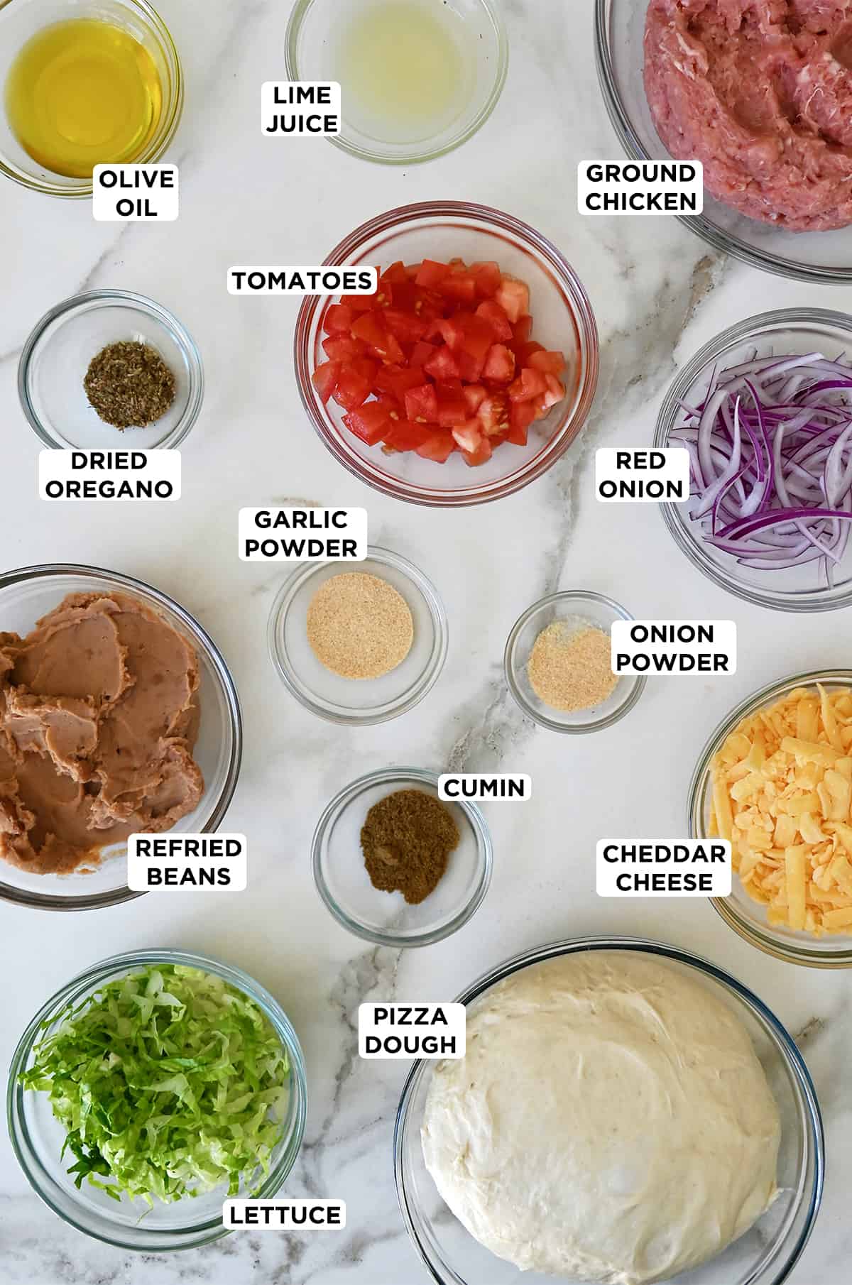Various sizes of glass bowls containing ingredients for taco pizza, including pizza dough, shredded lettuce, refried beans, dried oregano, diced tomatoes, ground chicken, sliced red onion, onion powder, garlic powder, cumin, and shredded cheddar cheese.