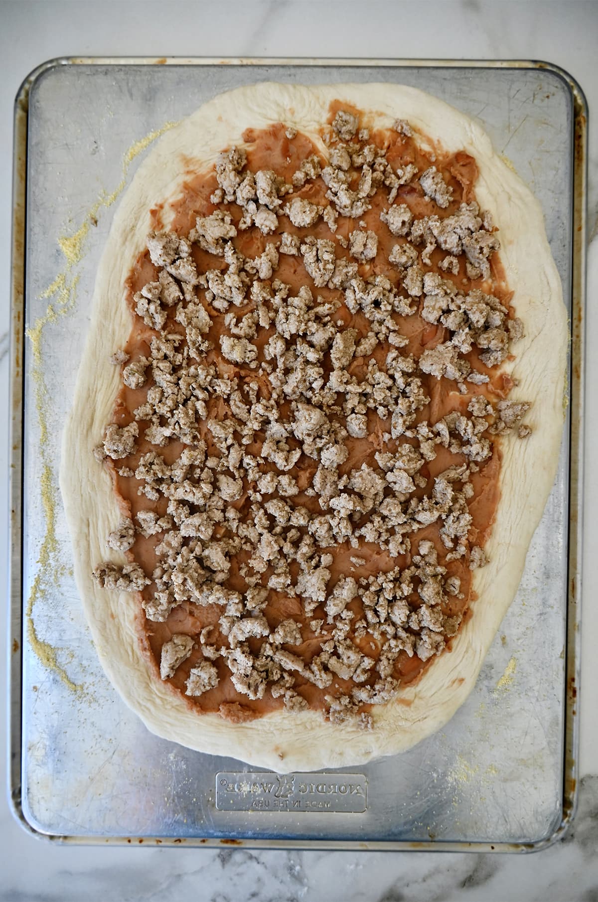 Pizza dough topped with refried beans and cooked ground chicken on a baking sheet.