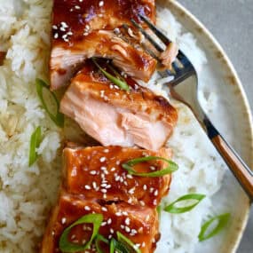 Baked honey garlic soy salmon garnished with sesame seeds and chopped scallions atop a bed of white rice on a plate with a fork.