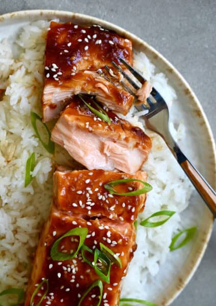 Baked honey garlic soy salmon garnished with sesame seeds and chopped scallions atop a bed of white rice on a plate with a fork.