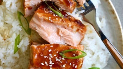 Baked honey garlic soy salmon garnished with sesame seeds and chopped scallions atop a bed of white rice on a plate with a fork.