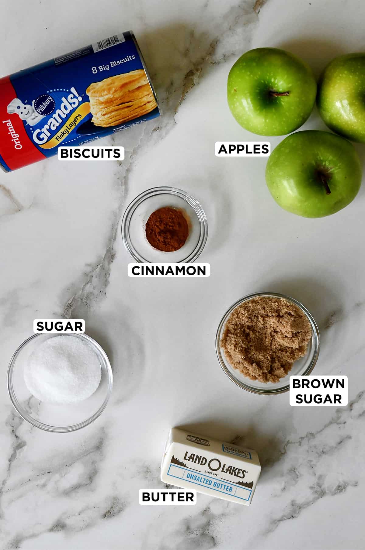 Pillsbury refrigerated biscuits next to Granny Smith apples and clear bowls containing brown sugar, ground cinnamon, sugar and butter.