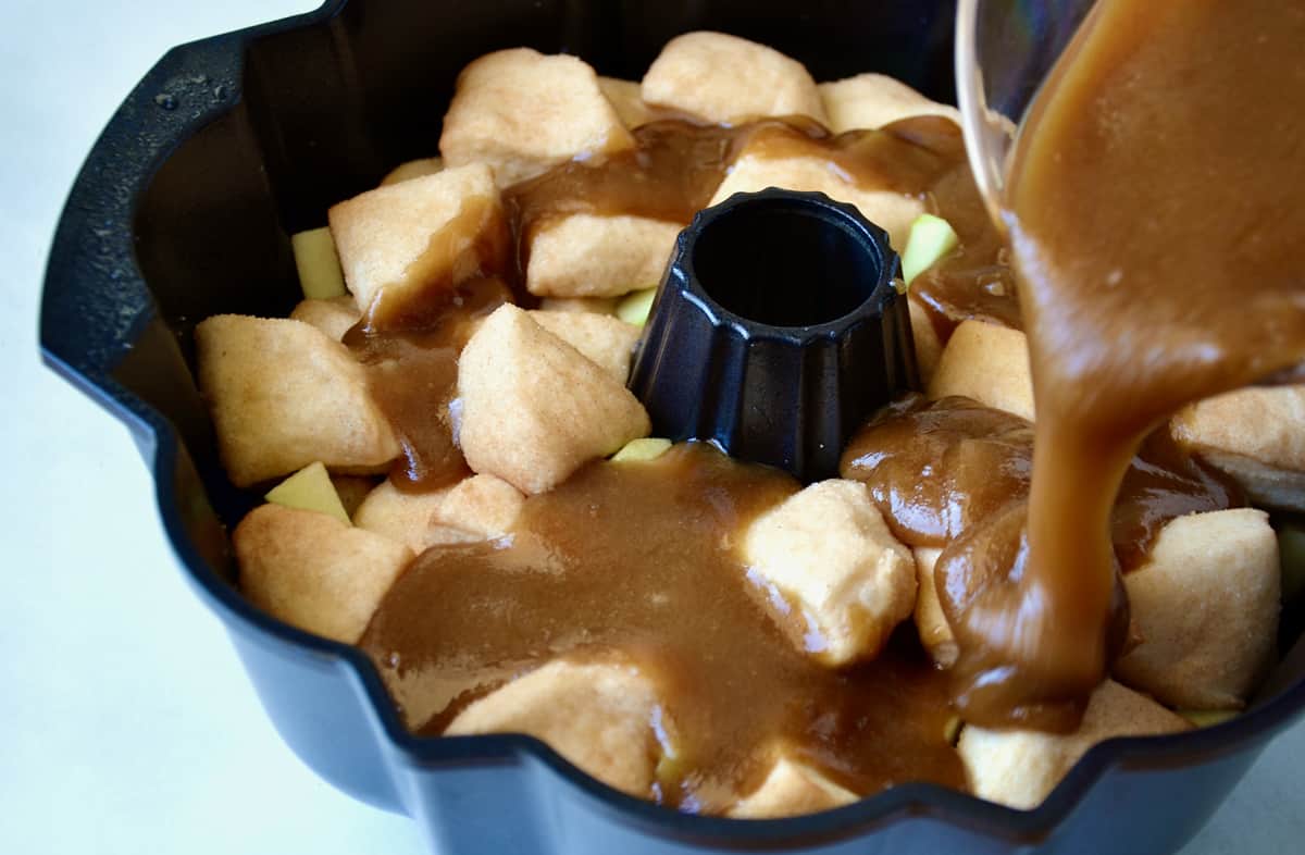 Caramel sauce being poured over biscuit dough pieces and diced green apples in a bundt pan.