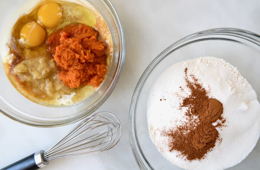 A whisk next to a clear bowl containing eggs, mashed fruit, and puree next to another clear bowl containing flour, sugar, and cinnamon