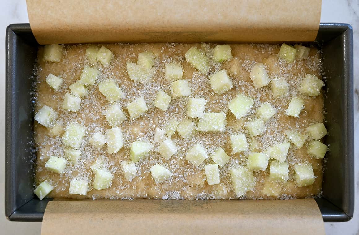 A top-down view of batter topped with diced apples in a parchment paper-lined loaf pan