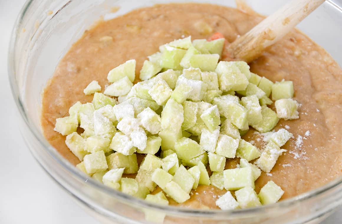 A spatula rests in a clear bowl containing batter and diced apples