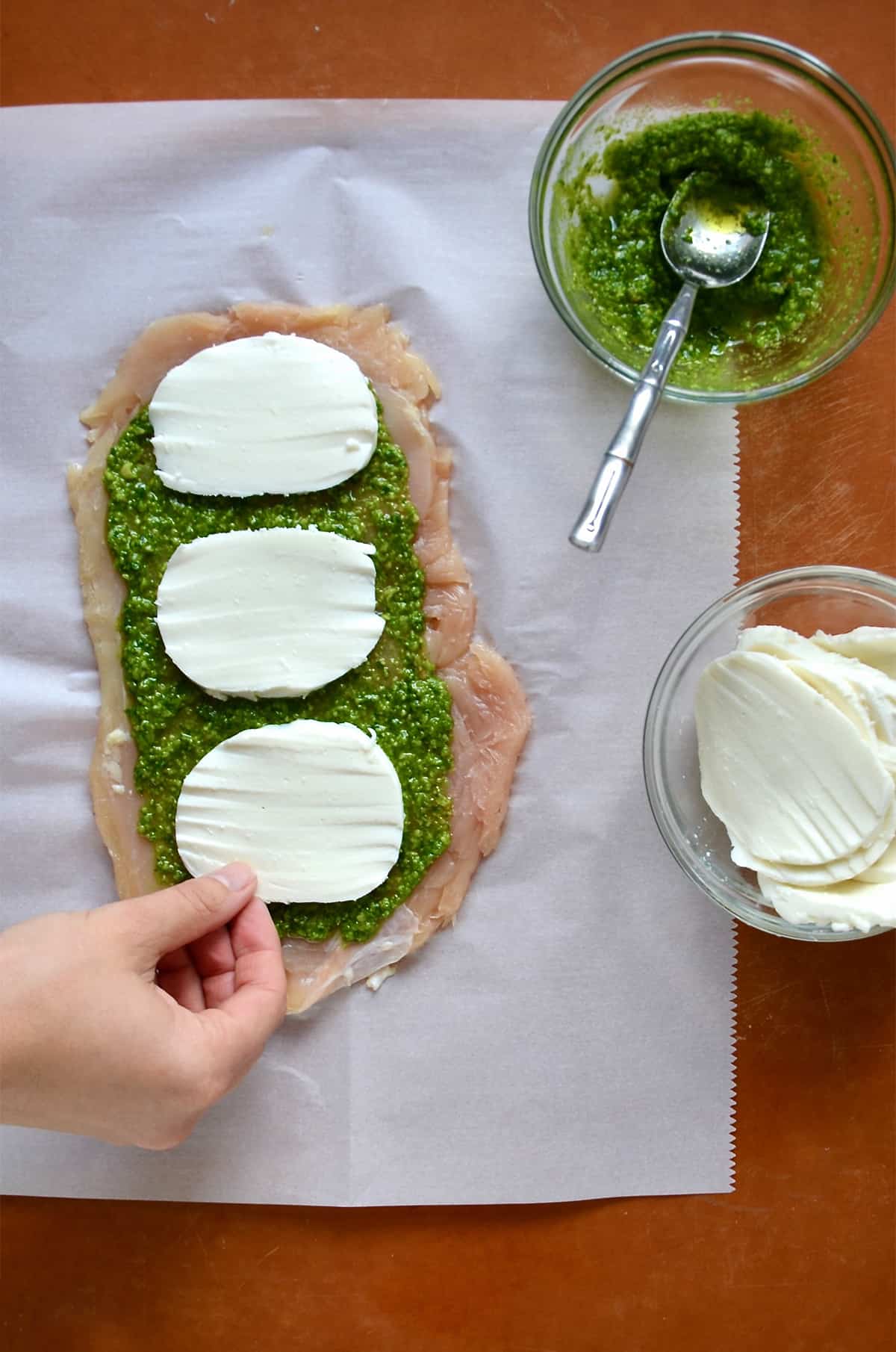 Thinly pounded chicken is topped with pesto and sliced mozzarella. Nearby are bowls of pesto and mozzarella cheese.