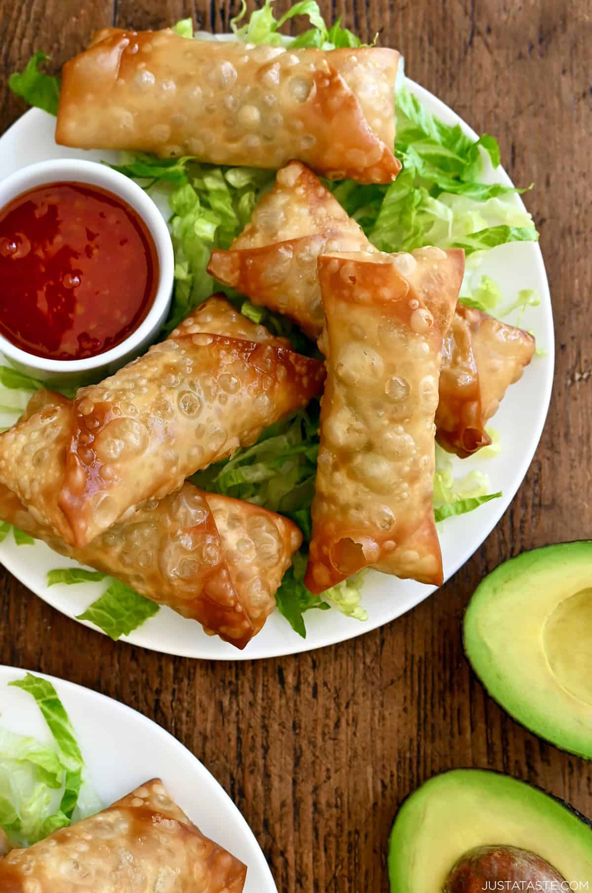 Extra-crispy avocado egg rolls atop a bed of shredded lettuce on a plate with a small bowl filled with Thai sweet chili sauce.