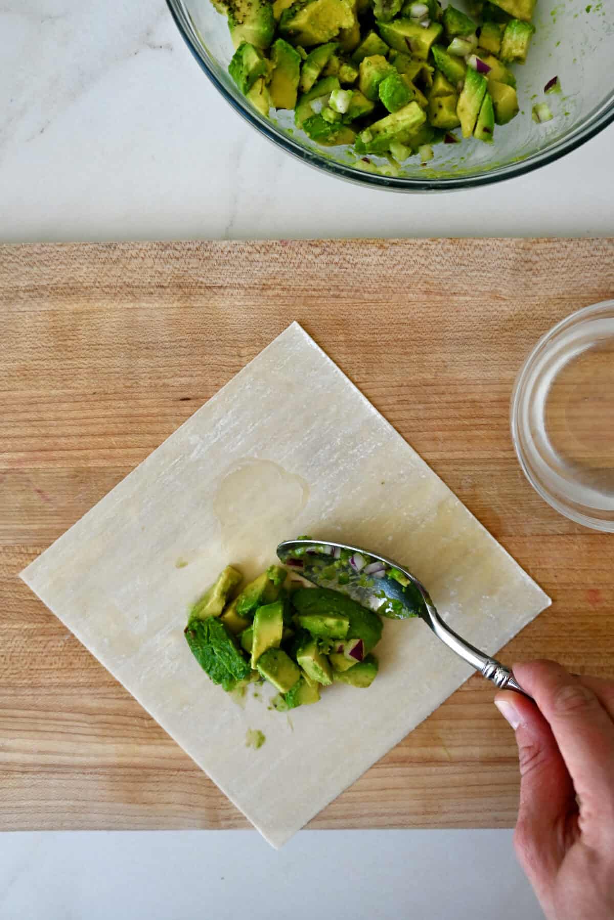 Avocado filling being spooned atop an egg roll wrapper that's on a wood cutting board along with a small glass bowl filled with water.