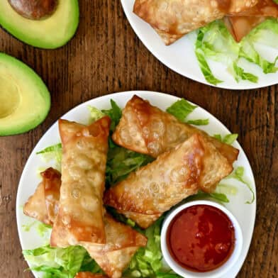 Avocado egg rolls atop shredded lettuce on a plate with a small bowl containing sweet chili sauce.