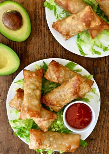 Avocado egg rolls atop shredded lettuce on a plate with a small bowl containing sweet chili sauce.