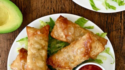 Avocado egg rolls atop shredded lettuce on a plate with a small bowl containing sweet chili sauce.