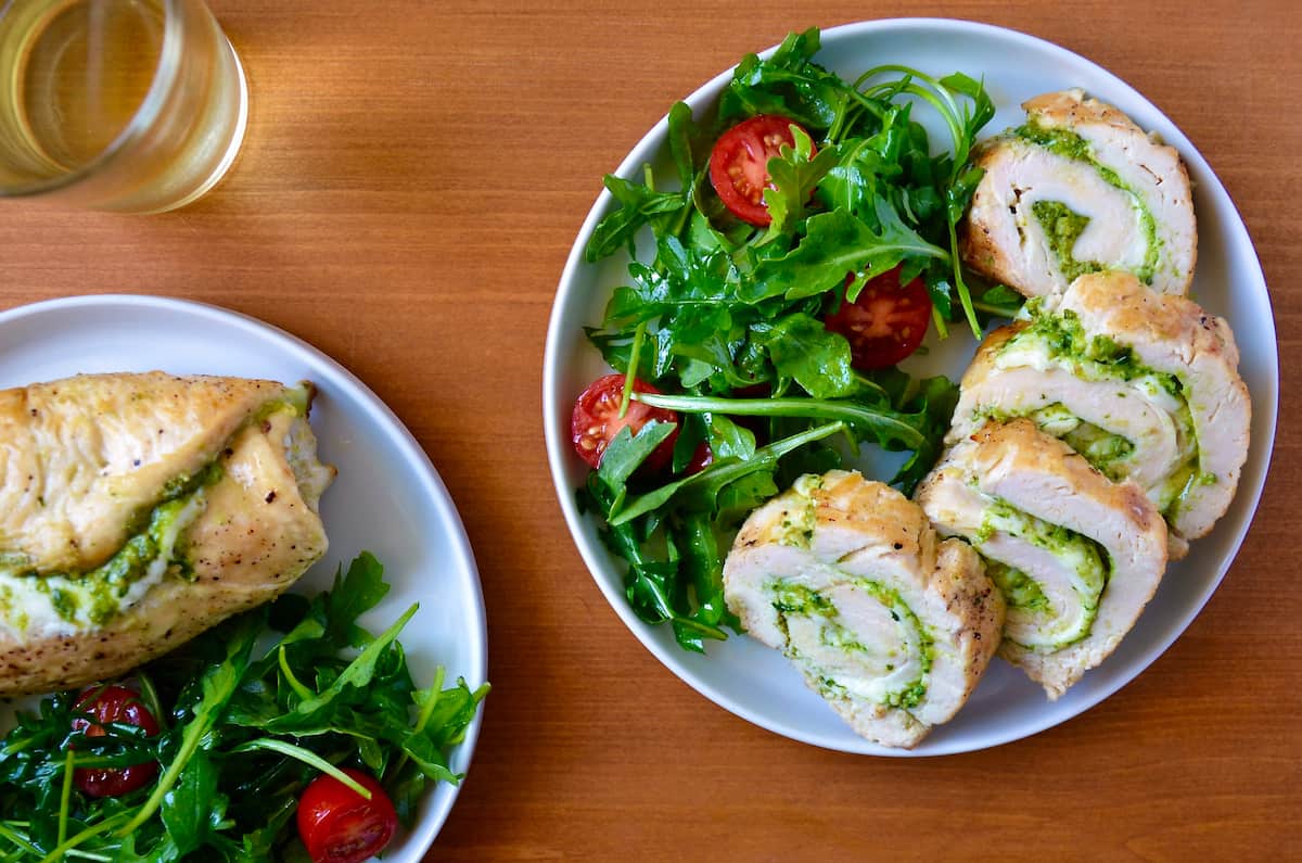 Sliced chicken roulades filled with pesto are on a plate with a salad of greens and tomatoes.